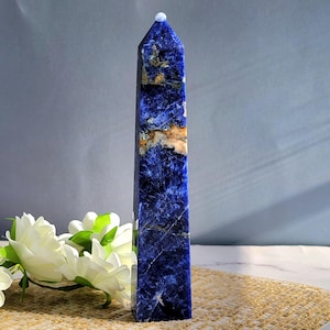 May include: A tall, rectangular sodalite obelisk with a polished finish. The stone is a deep blue with white and tan veining. A small, white, rounded stone sits atop the obelisk. The obelisk is displayed on a woven mat with white flowers.