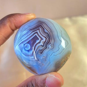 Puede incluir: Una piedra de &aacute;gata pulida y redondeada con intrincadas bandas conc&eacute;ntricas de color azul claro, blanco y violeta intenso. La superficie es lisa y reflectante, mostrando un patr&oacute;n natural en capas que irradia desde un punto central.