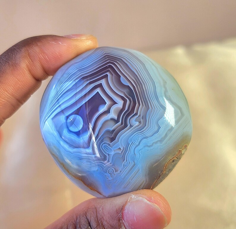 Puede incluir: Una piedra de &aacute;gata pulida y redondeada con intrincadas bandas conc&eacute;ntricas de color azul claro, blanco y violeta intenso. La superficie es lisa y reflectante, mostrando un patr&oacute;n natural en capas que irradia desde un punto central.