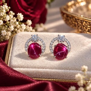 May include: A pair of stud earrings featuring oval-shaped deep red star gemstones set in silver-toned metal. Each earring is accented with a circular halo of small clear stones and a single larger clear stone at the top of the setting.