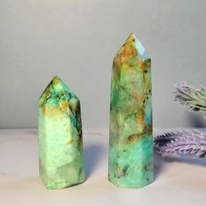 May include: Two polished crystal towers with a hexagonal shape. The stones feature a vibrant mix of mint green, translucent white, and warm amber-colored inclusions with dark mineral veins throughout the smooth, faceted surfaces of the points.
