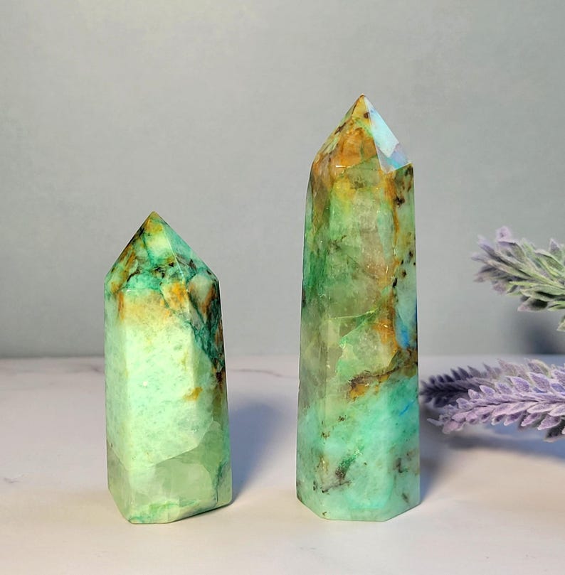 May include: Two polished crystal towers with a hexagonal shape. The stones feature a vibrant mix of mint green, translucent white, and warm amber-colored inclusions with dark mineral veins throughout the smooth, faceted surfaces of the points.