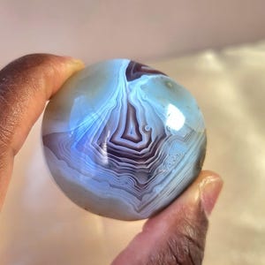 Puede incluir: Una piedra de &aacute;gata esf&eacute;rica y pulida que presenta intrincadas bandas conc&eacute;ntricas en tonos azul claro transl&uacute;cido, blanco y violeta intenso. La superficie lisa y redondeada muestra un patr&oacute;n natural en capas con un acabado brillante.