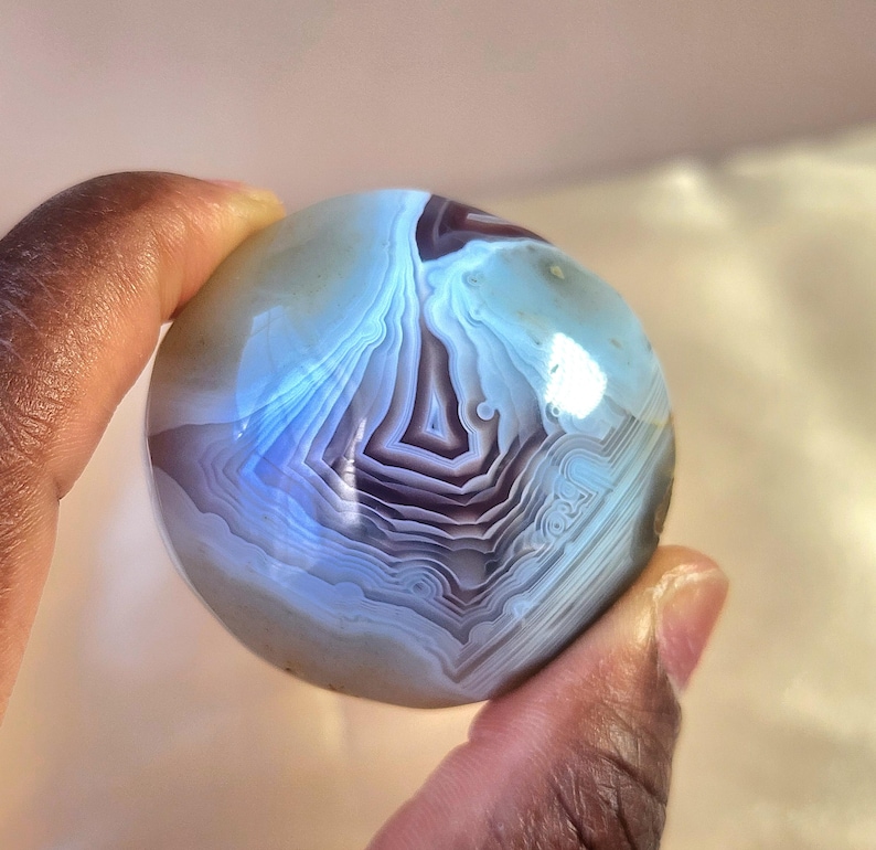 Puede incluir: Una piedra de &aacute;gata esf&eacute;rica y pulida que presenta intrincadas bandas conc&eacute;ntricas en tonos azul claro transl&uacute;cido, blanco y violeta intenso. La superficie lisa y redondeada muestra un patr&oacute;n natural en capas con un acabado brillante.