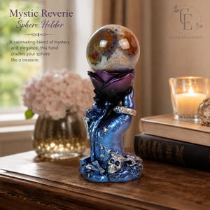 May include: A decorative sphere holder featuring a sculpted hand in a metallic blue finish, cradling a dark purple rose that supports a polished, multi-coloured agate sphere. The base includes a small skull and vine details with a silver ring.