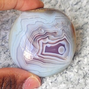 Puede incluir: Una piedra de &aacute;gata pulida y redondeada con bandas conc&eacute;ntricas de color azul claro, blanco y violeta suave. La superficie es lisa y reflectante, mostrando patrones intrincados y superpuestos que irradian desde el centro hacia los bordes.