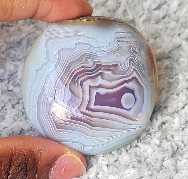 Puede incluir: Una piedra de &aacute;gata pulida y redondeada con bandas conc&eacute;ntricas de color azul claro, blanco y violeta suave. La superficie es lisa y reflectante, mostrando patrones intrincados y superpuestos que irradian desde el centro hacia los bordes.