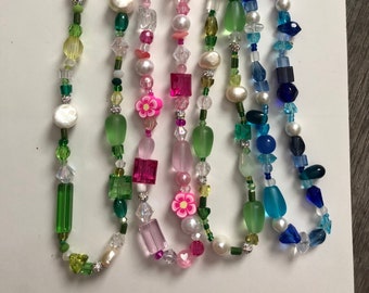 Op de afbeelding: Zes kralenkettingen met verschillende kleuren en vormen, waaronder groene, roze, blauwe, witte en heldere kralen. De kettingen hebben bloem- en vierkante kralen.