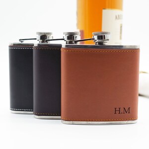 May include: Three leather-covered hip flasks with silver caps and a stitched design. The flask on the right has the initials "H.M." engraved on the front.