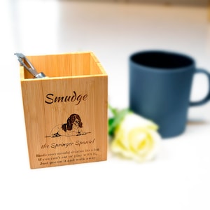 May include: A wooden pencil holder with a personalised inscription that reads "Smudge the Springer Spaniel". The inscription includes a quote about handling stressful situations like a dog. The holder is made of light brown bamboo and has a silver utensil inside.