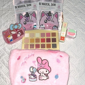 Puede incluir: Un surtido de productos de belleza que incluyen dos mascarillas faciales con diseños de unicornios, una paleta de sombras de ojos rosa, un neceser rosa de My Melody y otros dos artículos cosméticos. Las mascarillas dicen "BE MAGICAL, SKIN!"