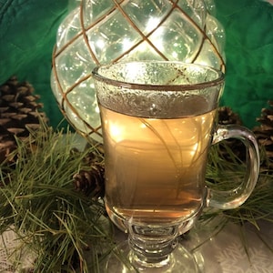 May include: A clear glass mug filled with a golden-colored liquid, possibly tea or a warm beverage. The mug sits on a reflective surface, surrounded by pine needles, pine cones, and a decorative glass orb with string lights. The background is a deep green.