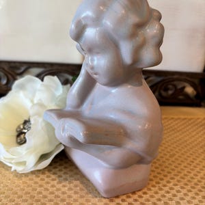 May include: A light pink ceramic figurine of a child reading a book. The figurine has a smooth, glossy finish with subtle blue and brown highlights. The child's face is turned downward, and the figurine is resting on a rectangular base.