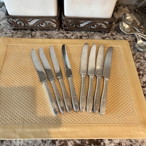 May include: A collection of eight silver butter knives arranged on a gold-colored placemat. The knives have rounded blades and ornate handles. Two white containers with decorative metal bases are in the background, along with a few silver spoons.