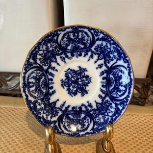 May include: A small, round, blue and white porcelain saucer with a gold rim. The saucer features a floral pattern in blue against a white background. The saucer is displayed on a gold-colored stand.