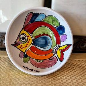 May include: A white ceramic bowl with a hand-painted fish design. The fish features vibrant colors including red, yellow, blue, green, and purple. The bowl has a simple, rounded shape and the word "CAMUSA" is written on the bottom.
