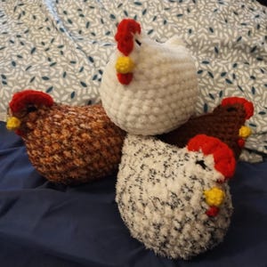 May include: Three crocheted chicken plushies. One is white with black speckles, one is brown with orange speckles, and one is brown with white speckles. All have red combs and yellow beaks.