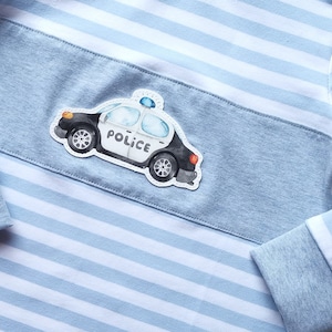 Könnte beinhalten: Hellblau-weiß gestreiftes Langarmshirt mit hellblauem Einsatz auf der Brust. Auf dem Einsatz ist ein Polizeiauto-Motiv zentriert. Das Shirt hat hellblaue Bündchen und einen hellblauen Kragen.