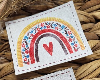 May include: Two white rectangular patches with a rainbow design. The rainbow features yellow, orange, red, and brown arches, adorned with floral patterns and a red heart. The patches are stitched with a dotted border.