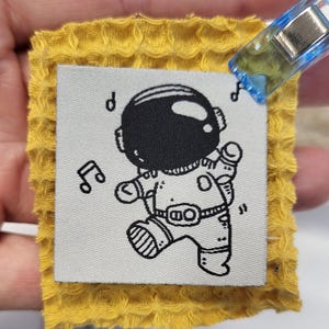 Può includere: Una piccola etichetta quadrata in tessuto con un astronauta dei cartoni animati che balla, stampata in nero su sfondo bianco. L'etichetta è attaccata a un tessuto giallo strutturato. Sull'etichetta sono stampate anche note musicali.