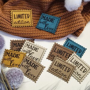 Op de afbeelding: Een verzameling van zes kleine, rechthoekige, bruine leren labels met witte stiksels. De labels hebben verschillende tekst, waaronder "LIMITED edition", "MADE with love" en "MADE with love". De labels zijn gerangschikt op een wit oppervlak met een grijs, pluche oppervlak in de rechterbenedenhoek.