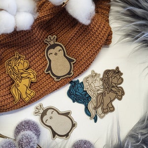 May include: Four felt patches featuring a penguin and a unicorn. The penguin is wearing a crown and has a closed-eye smile. The unicorn has a crown and a closed-eye smile. The patches are brown, tan, and blue.