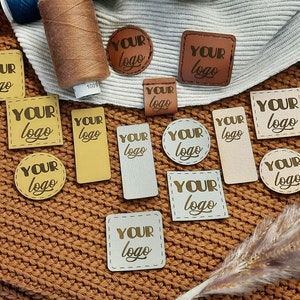 Puede incluir: Una colección de etiquetas de cuero marrón, beige y blanco con el texto "YOUR LOGO" en letras doradas. Las etiquetas están dispuestas sobre un fondo de tejido de punto marrón.
