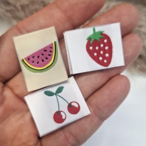 May include: Three fabric labels with fruit designs: a watermelon slice, a strawberry, and two cherries. The watermelon is pink with black seeds and a yellow rind. The strawberry is red with white seeds and a green stem. The cherries are red with green stems.