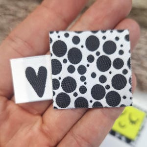May include: Close-up of three small fabric tags. One tag is white with a black heart, another is white with black polka dots, and the third is yellow with a black and white pattern and a smiling face.