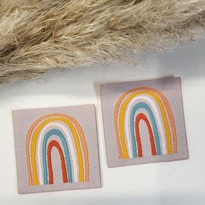 Puede incluir: Dos parches cuadrados de tela con diseños de arcoíris bordados. Cada arcoíris presenta arcos en naranja, amarillo, blanco, turquesa y rojo. Los parches están sobre un fondo gris claro. Hierba ornamental seca en el fondo.