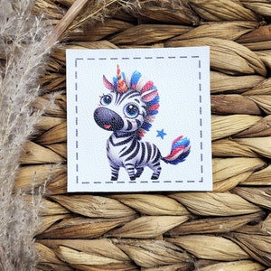 Op de afbeelding: Een vierkante patch met een cartoon zebra met een eenhoornhoorn en kleurrijke manen en staart. De zebra heeft zwart-witte strepen, grote blauwe ogen en een kleine blauwe ster. De patch staat op een getextureerde witte achtergrond.