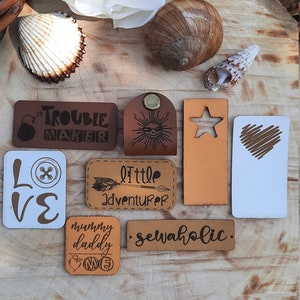 Op de afbeelding: Assortiment van leren labels met verschillende ontwerpen en tekst, waaronder "Trouble Maker", "Little Adventurer", "Sewahollic", "Mommy Daddy Me" en "Love". De labels liggen op een houten oppervlak.