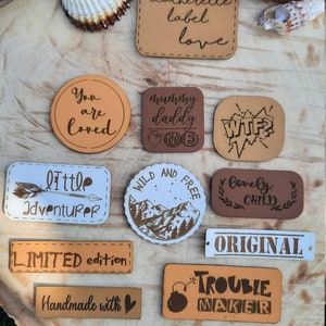 May include: A collection of leatherette labels with various phrases and designs. The labels are in different shapes and sizes, with text such as "love", "You are loved", "Little Adventurer", and "Trouble Maker".
