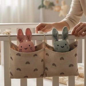 May include: Two fabric storage organizers hanging on a white crib. Each organizer has a pocket with a smiling bunny toy. One bunny is pink, the other is green. The organizers are beige with black rainbow patterns.