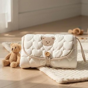 May include: Cream-coloured baby changing mat with a heart-shaped quilted pattern and a small teddy bear appliqué. The mat has a flap closure with a wooden button and a matching teddy bear plush toy.