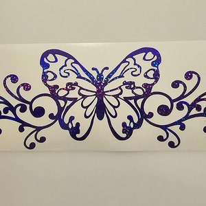 Puede incluir: Una mariposa de purpurina iridiscente morada con remolinos decorativos a cada lado. La mariposa est&aacute; delineada en negro y tiene un interior blanco.