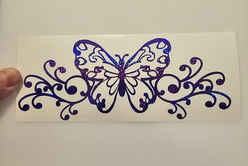 Puede incluir: Una mariposa de purpurina iridiscente morada con remolinos decorativos a cada lado. La mariposa est&aacute; delineada en negro y tiene un interior blanco.