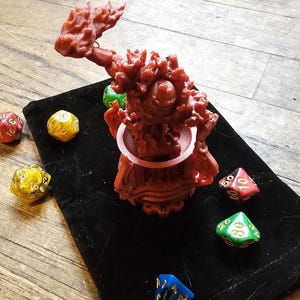 Puede incluir: Una miniatura roja impresa en 3D de una criatura con un brazo levantado sosteniendo un arma. La miniatura está sentada sobre una bolsa de terciopelo negro con varios dados de colores.