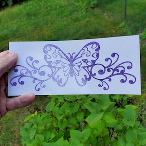 Puede incluir: Calcoman&iacute;a de vinilo con purpurina morada de una mariposa con enredaderas en espiral. La calcoman&iacute;a est&aacute; sobre un fondo blanco.