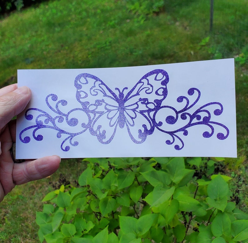 Puede incluir: Calcoman&iacute;a de vinilo con purpurina morada de una mariposa con enredaderas en espiral. La calcoman&iacute;a est&aacute; sobre un fondo blanco.