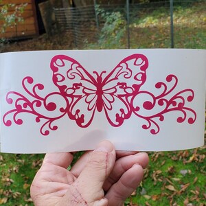 Puede incluir: Una mariposa rosa con intrincados detalles en las alas y un dise&ntilde;o floral a cada lado. La mariposa y el dise&ntilde;o floral est&aacute;n cortados en vinilo y est&aacute;n listos para ser aplicados a una superficie.