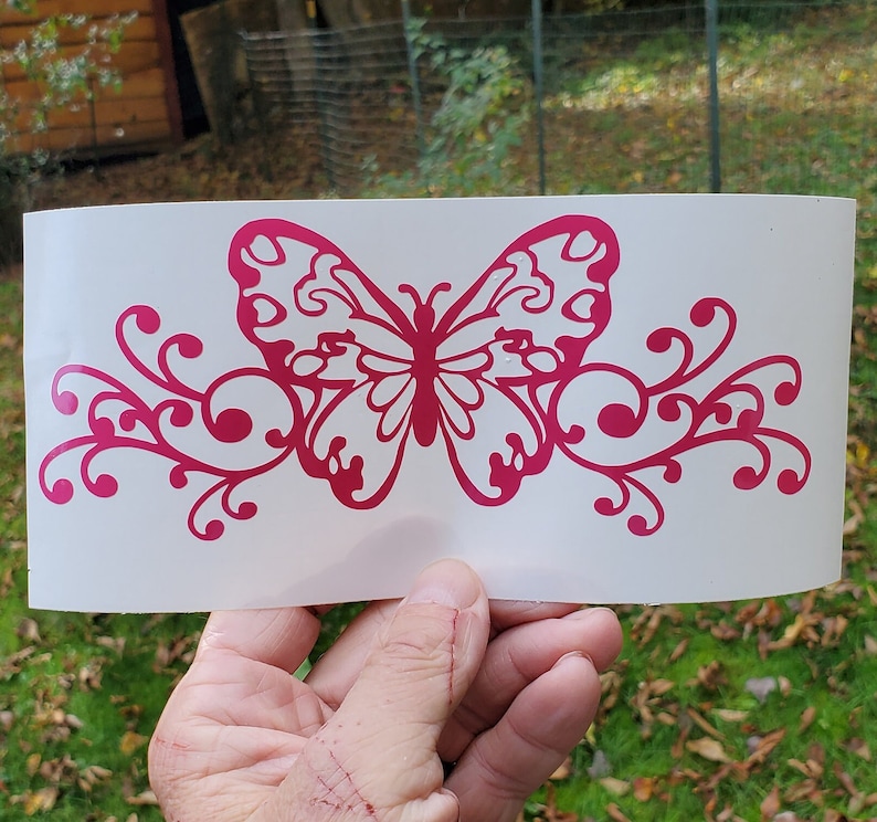 Puede incluir: Una mariposa rosa con intrincados detalles en las alas y un dise&ntilde;o floral a cada lado. La mariposa y el dise&ntilde;o floral est&aacute;n cortados en vinilo y est&aacute;n listos para ser aplicados a una superficie.