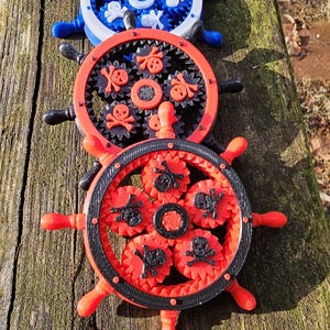 Può includere: Tre timoni decorativi blu, rossi e neri, con intricati disegni di ingranaggi e teschi. Le ruote sono disposte su una superficie di legno stagionato, che mostra un tema nautico o pirata.