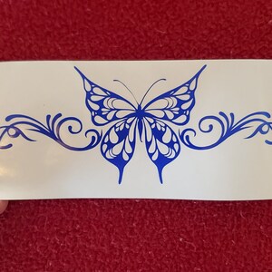 May include: A blue butterfly with intricate wing patterns is centered on a white background. The butterfly is surrounded by a decorative, swirling design.