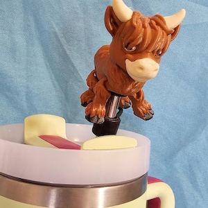 Puede incluir: Un tapón de botella de vaca Highland marrón y blanco con textura peluda. El tapón está unido a una base de metal negro y está sentado encima de una taza de viaje amarilla y blanca.