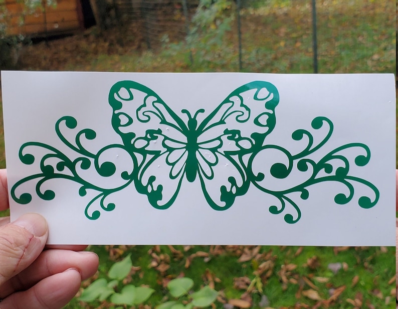 Puede incluir: Calcoman&iacute;a de vinilo verde de una mariposa con acentos florales en espiral. La mariposa tiene un dise&ntilde;o detallado con alas intrincadas.