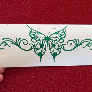 May include: A green butterfly with white accents and intricate wing details is centered on a white background. The butterfly is surrounded by a decorative, swirling vine design.