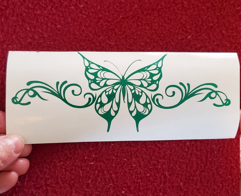 May include: A green butterfly with white accents and intricate wing details is centered on a white background. The butterfly is surrounded by a decorative, swirling vine design.
