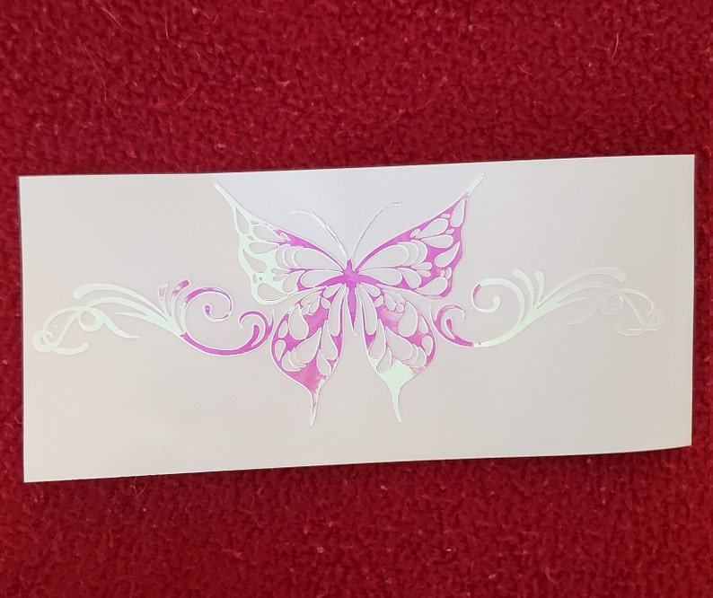 May include: A white vinyl decal with a pink butterfly design. The butterfly has intricate details and is surrounded by a decorative scrollwork design.