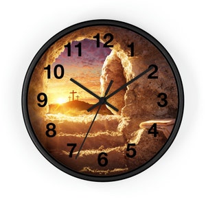 May include: A round clock with a black frame and a sunrise scene with a stone tomb and three crosses. The clock face has Roman numerals and black hands.